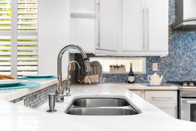 details with white cabinets, wall chimney range hood, sink, and tasteful backsplash