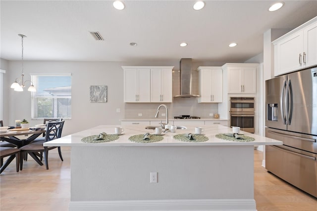 kitchen featuring decorative light fixtures, a center island with sink, stainless steel appliances, and wall chimney range hood