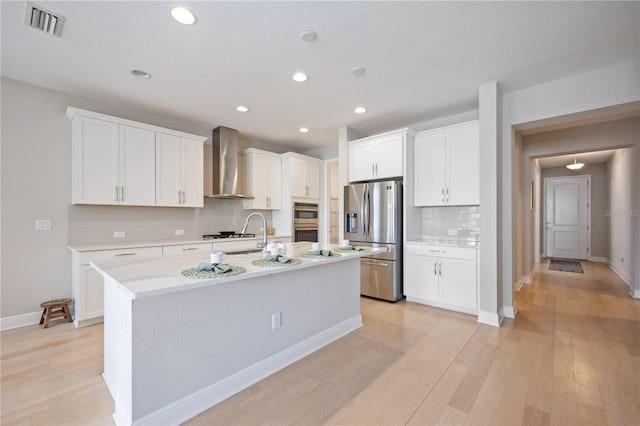 kitchen with backsplash, stainless steel appliances, wall chimney range hood, white cabinetry, and an island with sink