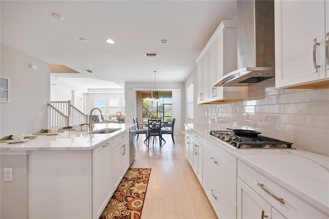 kitchen featuring wall chimney range hood, sink, an island with sink, light stone counters, and white cabinetry