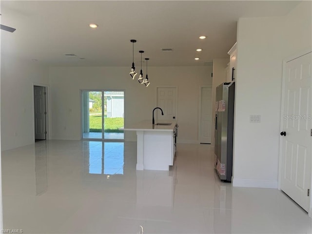 kitchen with a center island with sink, stainless steel fridge, sink, white cabinetry, and decorative light fixtures