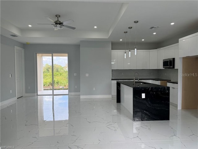 kitchen featuring a kitchen island with sink, a raised ceiling, hanging light fixtures, sink, and white cabinetry