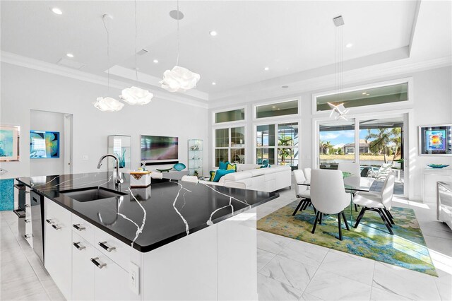 kitchen with a tray ceiling, a kitchen island with sink, sink, decorative light fixtures, and white cabinetry