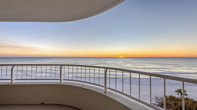 balcony at dusk featuring a beach view and a water view