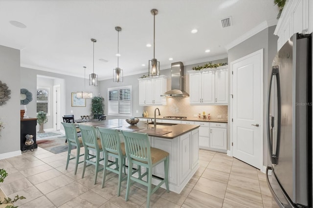 kitchen featuring stainless steel appliances, a kitchen island with sink, wall chimney range hood, white cabinetry, and hanging light fixtures