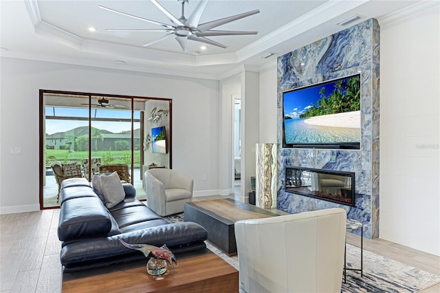 living room with a high end fireplace, a tray ceiling, ceiling fan, crown molding, and wood-type flooring