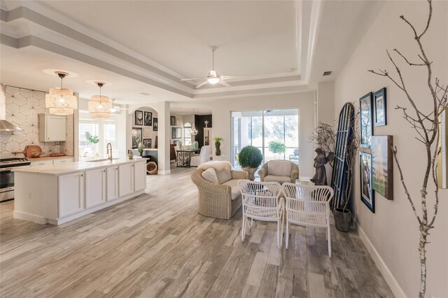 living room with a raised ceiling, sink, ceiling fan with notable chandelier, and light wood-type flooring