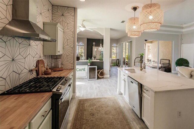 kitchen with wall chimney range hood, sink, hanging light fixtures, appliances with stainless steel finishes, and light hardwood / wood-style floors