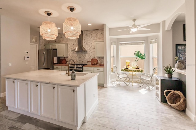 kitchen featuring white cabinets, wall chimney exhaust hood, an island with sink, appliances with stainless steel finishes, and decorative light fixtures