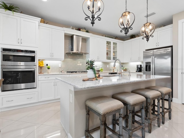 kitchen featuring a center island with sink, stainless steel appliances, glass insert cabinets, white cabinets, and wall chimney exhaust hood