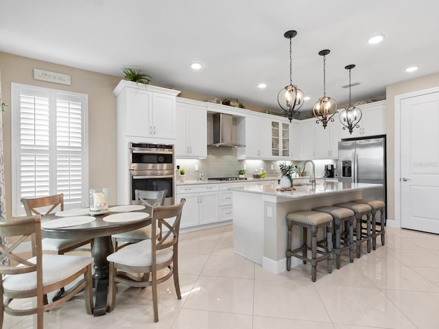 kitchen with glass insert cabinets, appliances with stainless steel finishes, white cabinets, an island with sink, and wall chimney exhaust hood