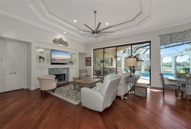 living room with ceiling fan, dark wood-type flooring, a premium fireplace, a tray ceiling, and ornamental molding