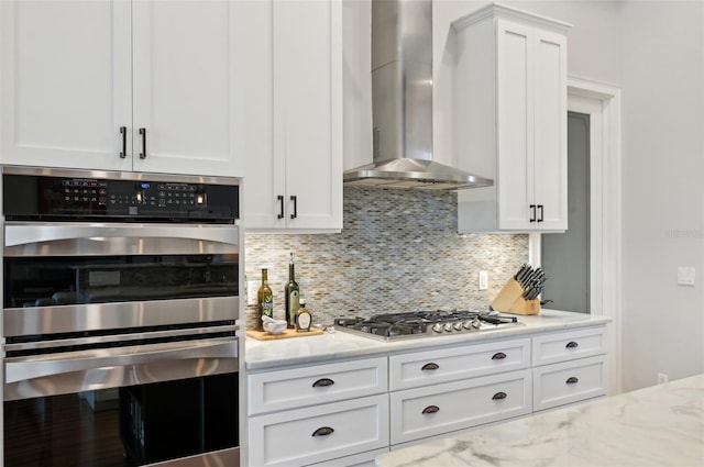 kitchen with light stone countertops, wall chimney exhaust hood, white cabinetry, stainless steel appliances, and decorative backsplash