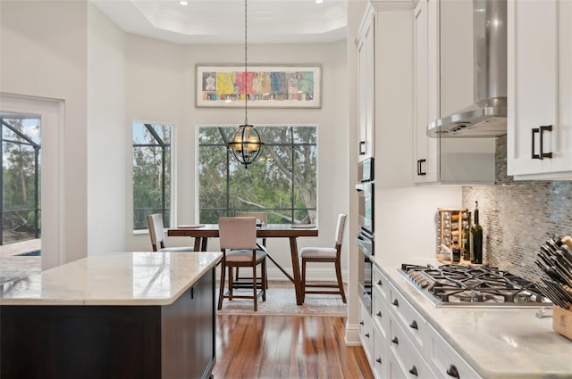 kitchen featuring pendant lighting, wall chimney range hood, white cabinetry, a notable chandelier, and light stone counters