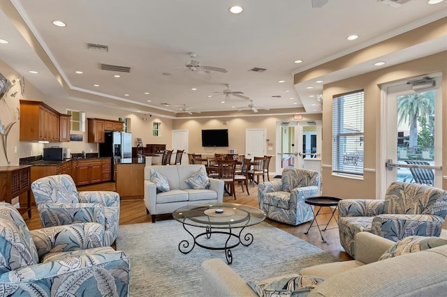 living room with light wood-type flooring, a raised ceiling, ceiling fan, and ornamental molding