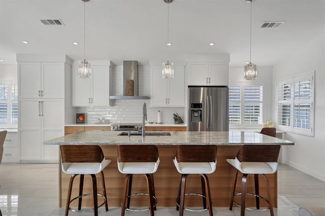 kitchen with stainless steel refrigerator with ice dispenser, an island with sink, white cabinetry, and wall chimney range hood