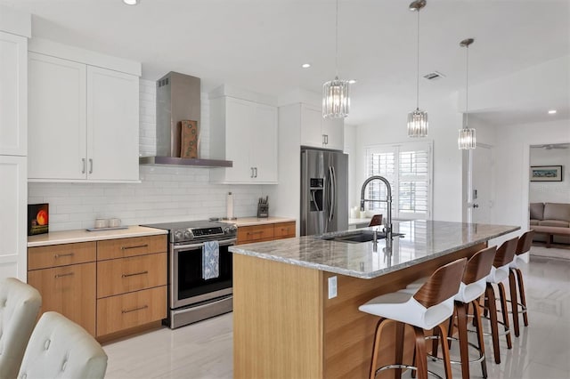 kitchen with sink, stainless steel appliances, wall chimney range hood, an island with sink, and white cabinets