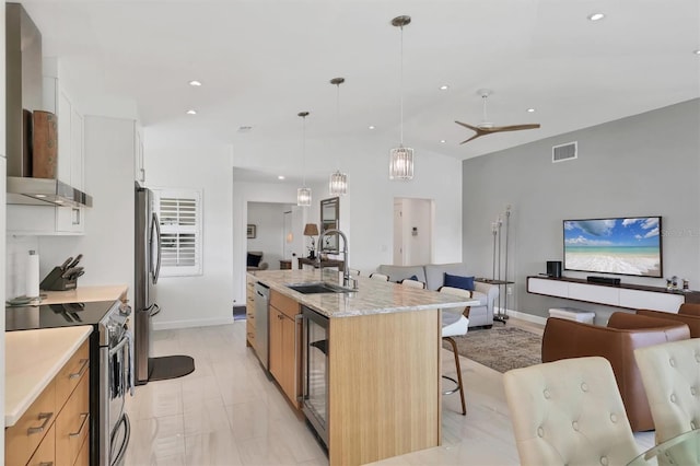 kitchen with wall chimney range hood, sink, an island with sink, appliances with stainless steel finishes, and light stone counters