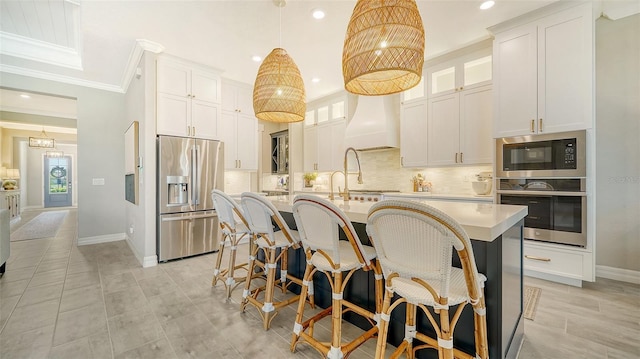 kitchen with pendant lighting, custom range hood, appliances with stainless steel finishes, white cabinetry, and a center island with sink