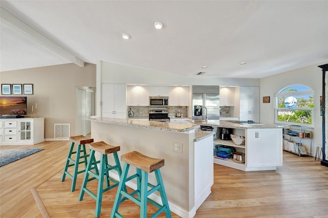 kitchen with light stone countertops, lofted ceiling with beams, a kitchen island with sink, white cabinets, and appliances with stainless steel finishes