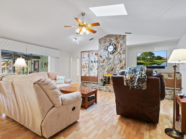 living room with ceiling fan, a stone fireplace, light wood-type flooring, and lofted ceiling with skylight