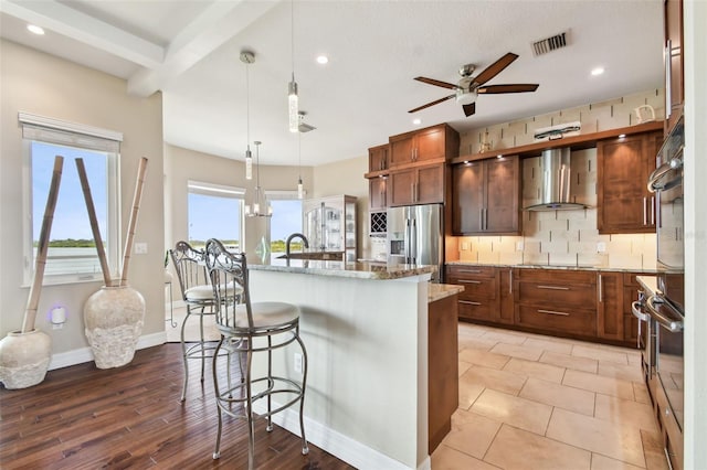 kitchen with light stone countertops, wall chimney exhaust hood, hanging light fixtures, stainless steel fridge with ice dispenser, and a kitchen island with sink