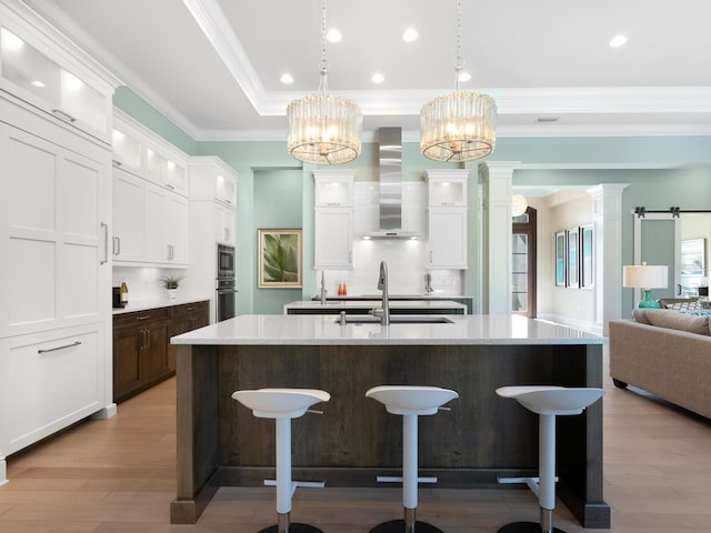 kitchen featuring wall chimney range hood, sink, a center island with sink, white cabinets, and hanging light fixtures