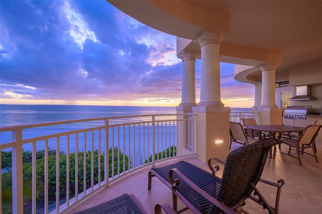 balcony at dusk featuring area for grilling, a water view, and a beach view