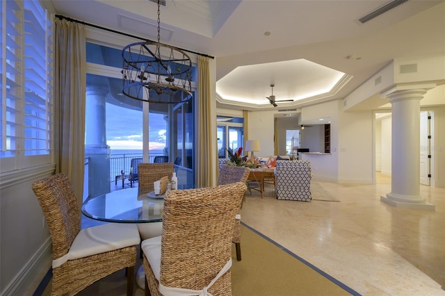 dining area with a tray ceiling, ornate columns, a water view, and ceiling fan with notable chandelier