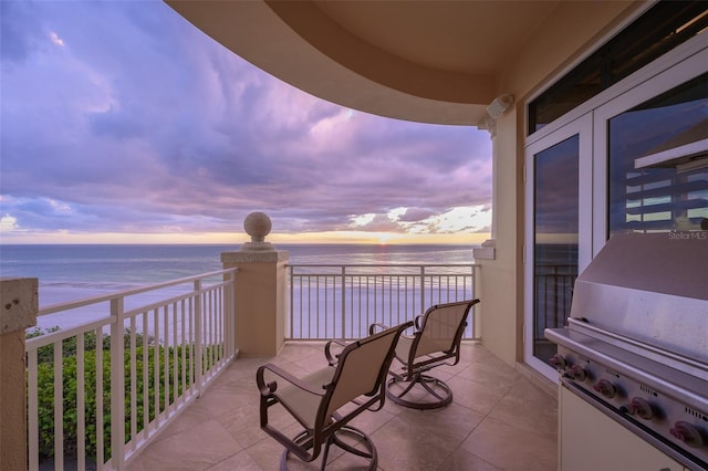 balcony at dusk with a view of the beach, a water view, and grilling area