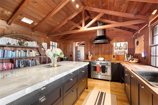 kitchen with wall chimney exhaust hood, lofted ceiling with beams, light hardwood / wood-style flooring, stainless steel stove, and wood walls