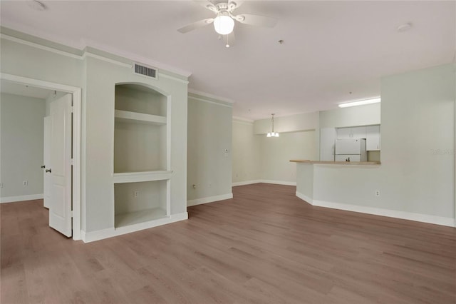 unfurnished living room with crown molding, built in shelves, wood-type flooring, and ceiling fan with notable chandelier