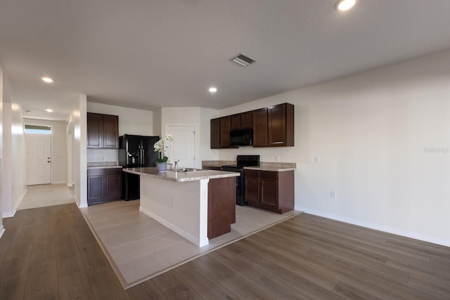 kitchen featuring sink, a kitchen island with sink, dark brown cabinetry, black appliances, and light hardwood / wood-style floors