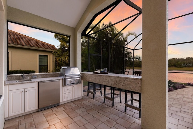 patio terrace at dusk with glass enclosure, a grill, an outdoor wet bar, and an outdoor kitchen