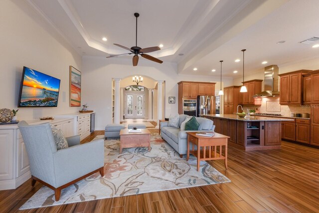 living room with decorative columns, light wood-type flooring, a tray ceiling, ceiling fan with notable chandelier, and ornamental molding