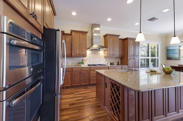 kitchen featuring wall chimney exhaust hood, stainless steel appliances, a kitchen island with sink, sink, and decorative light fixtures