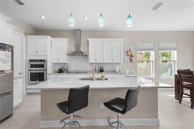 kitchen featuring a kitchen island with sink, wall chimney exhaust hood, hanging light fixtures, and appliances with stainless steel finishes