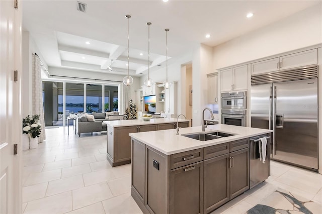 kitchen featuring a kitchen island with sink, coffered ceiling, sink, appliances with stainless steel finishes, and decorative light fixtures