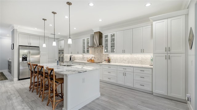 kitchen with light stone countertops, wall chimney range hood, an island with sink, white cabinets, and appliances with stainless steel finishes