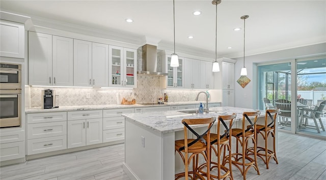 kitchen with sink, wall chimney range hood, decorative light fixtures, a center island with sink, and white cabinets