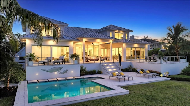 back house at dusk featuring a balcony, an outdoor living space, ceiling fan, a patio area, and a lawn