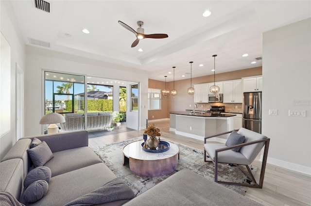 living room with ceiling fan with notable chandelier, light hardwood / wood-style floors, sink, and a tray ceiling