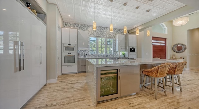 kitchen featuring white cabinets, hanging light fixtures, wine cooler, an island with sink, and double oven