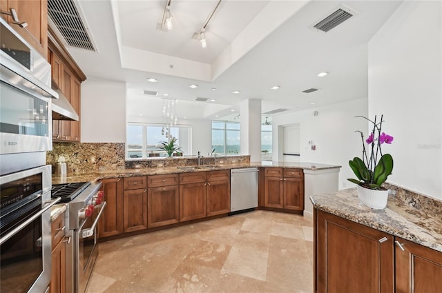 kitchen with appliances with stainless steel finishes, a sink, brown cabinets, light stone countertops, and a raised ceiling