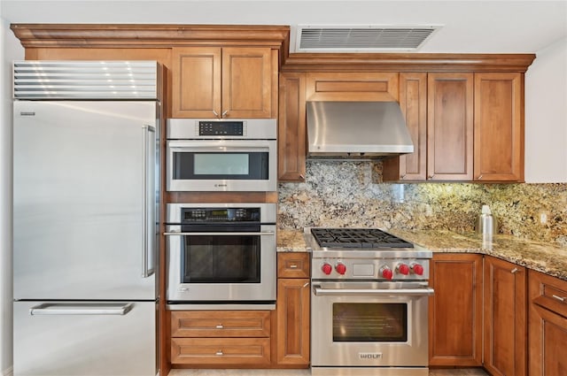 kitchen with high quality appliances, wall chimney exhaust hood, brown cabinetry, and tasteful backsplash