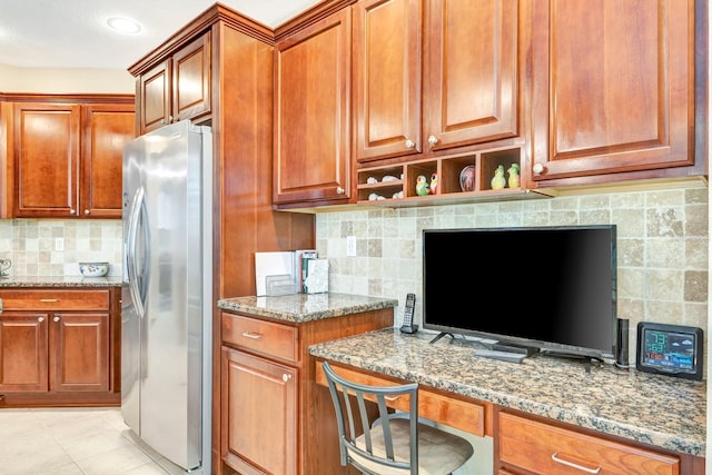 kitchen featuring light stone countertops, stainless steel refrigerator with ice dispenser, backsplash, light tile patterned floors, and built in desk