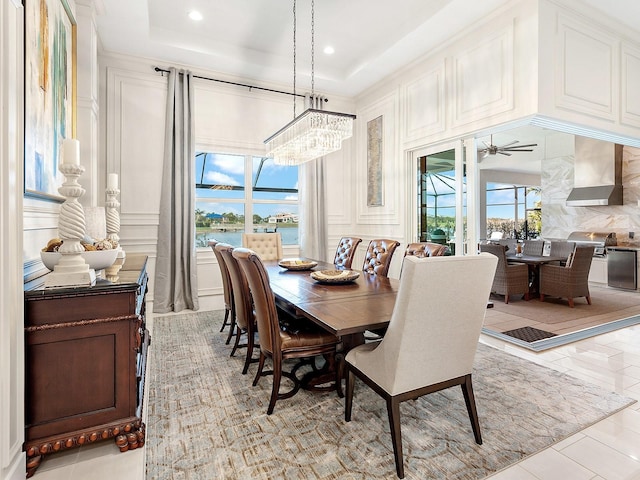 tiled dining space with ceiling fan with notable chandelier, a healthy amount of sunlight, a water view, and a tray ceiling