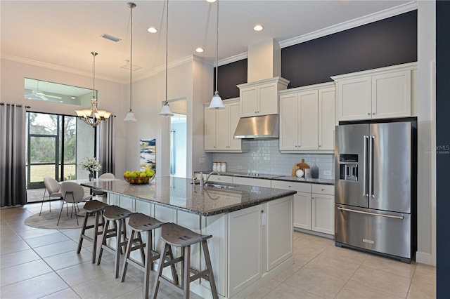 kitchen with high end fridge, white cabinetry, under cabinet range hood, and an island with sink