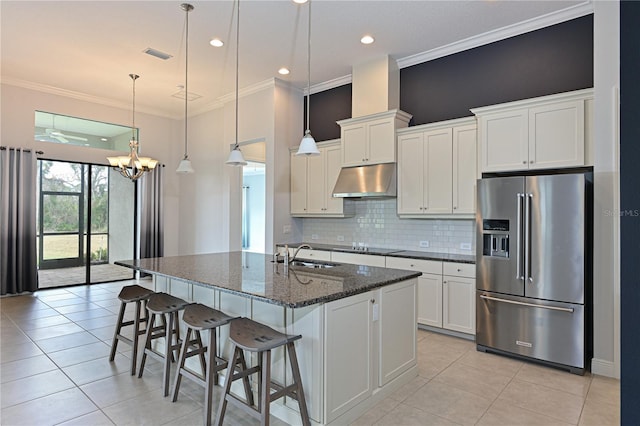 kitchen featuring a kitchen island with sink, under cabinet range hood, white cabinetry, high end fridge, and dark stone counters