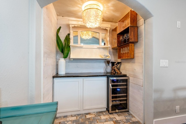 bar featuring wood walls, white cabinets, dark stone countertops, beverage cooler, and a chandelier